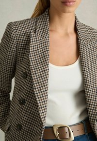 Smeđi houndstooth sako s crnim dugmadima, šiljatim reverima, nosi se preko bijelog rebrastog topa i smeđeg remena sa zlatnom kopčom.