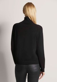 Blonde vrouw die met haar rug naar de camera staat, gekleed in een zwarte coltrui en zwarte leren broek, tegen een effen lichte achtergrond.
