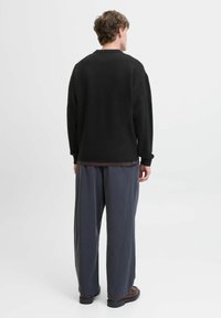Svart strikket genser med rund hals og ribbestrikket kant, kombinert med løstsittende grå bukser og brune sko. Uformell design, teksturert stoff.