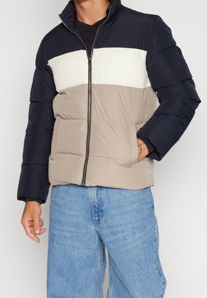 Homme portant une doudoune à blocs de couleurs avec des sections bleu marine, blanc et beige, un jean bleu, et une main dans la poche de la veste.