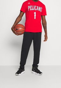 Red short-sleeve t-shirt with "PELICANS" and number 1, paired with black sweatpants and black athletic shoes; holds a basketball.