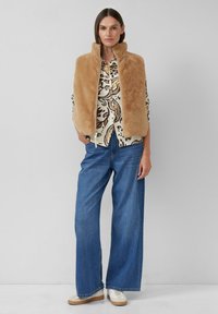 Vrouw in een beige nep-vacht vest, met een gebloemdblouse, wijde blauwe jeans en witte sneakers, staande tegen een effen lichte achtergrond.