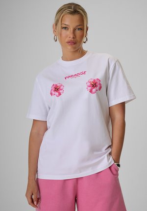 Kobieta ubrana w biały t-shirt z różowymi kwiatami i tekstem "Paradise", w połączeniu z luźnymi różowymi szortami, stojąca na jednolitym szarym tle.