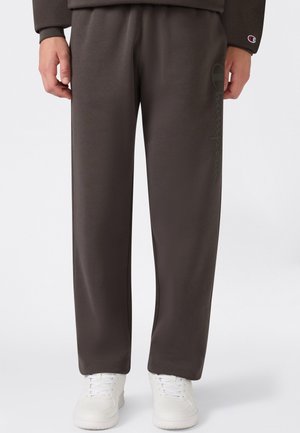 Personne portant un pantalon ample gris foncé avec un logo vertical subtil sur la jambe gauche et des baskets blanches, debout devant un fond uni.