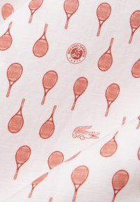 Tessuto bianco decorato con motivo a racchette da tennis arancioni ripetute, un piccolo logo di coccodrillo e un emblema circolare "Roland Garros Paris".