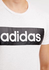 Camiseta de algodón blanca con cuello redondo, que presenta un gráfico rectangular negro destacado con el texto blanco "adidas" centrado en el pecho.