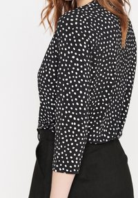 Czarna bluzka w białe kropki, z długimi rękawami i zaokrąglonym dekoltem. Materiał wydaje się lekki o gładkiej fakturze.