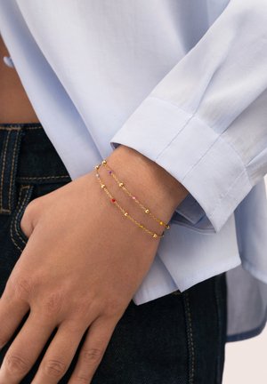 Pulsera de doble cadena dorada con pequeñas cuentas de colores en la muñeca, llevada con una camisa de manga larga azul claro y vaqueros oscuros.