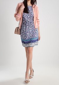 Vestido con estampado floral sobre una base blanca, con detalles en azul y verde, cintura ceñida, combinado con una chaqueta bomber rosa y sandalias de tacón grises.