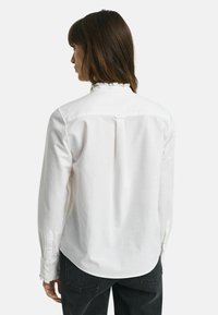 Camicia bianca con colletto arricciato e maniche lunghe, dotata di una piega sul retro e di una texture sottile. Abbinata a jeans di denim scuri.