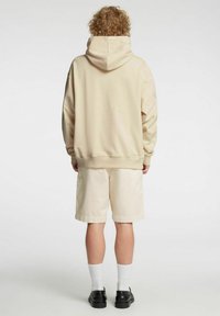 Beige hettegenser med hette, kombinert med kremfargede shorts. Mykt bomullsstoff, avslappet passform. Svarte sko og hvite sokker fullfører antrekket.