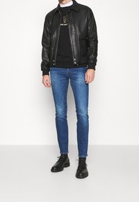 Uomo che indossa una giacca di pelle nera, una felpa nera con logo dorato, jeans slim fit blu e stivali neri con lacci, in piedi su uno sfondo bianco.