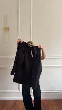 Črna srajca s knofki, dolgimi rokavi, ovratnikom in sprednjim žepom. Gladek tekstura in raven kroj, prikazana na enobarvnem ozadju.