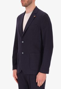 Blazer blu con texture, colletto a taglio, due bottoni e tasche frontali, indossato su una camicia chiara. Presenta un pin decorativo sul colletto.