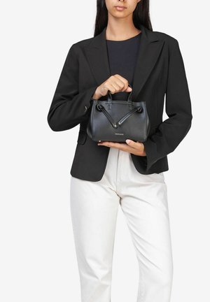 Bolso de cuero negro con asas cortas, forma estructurada y acentos dorados. Una mujer lo sostiene mientras viste un blazer negro y pantalones blancos.