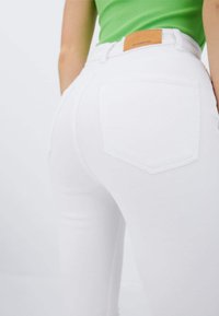 Jean en denim blanc avec une coupe ajustée, avec une poche arrière et une étiquette en cuir marron sur la taille. Texture lisse et design épuré.