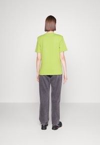 Camiseta verde de manga corta hecha de algodón, combinada con pantalones de pierna ancha grises, mostrada desde atrás sobre un fondo liso.