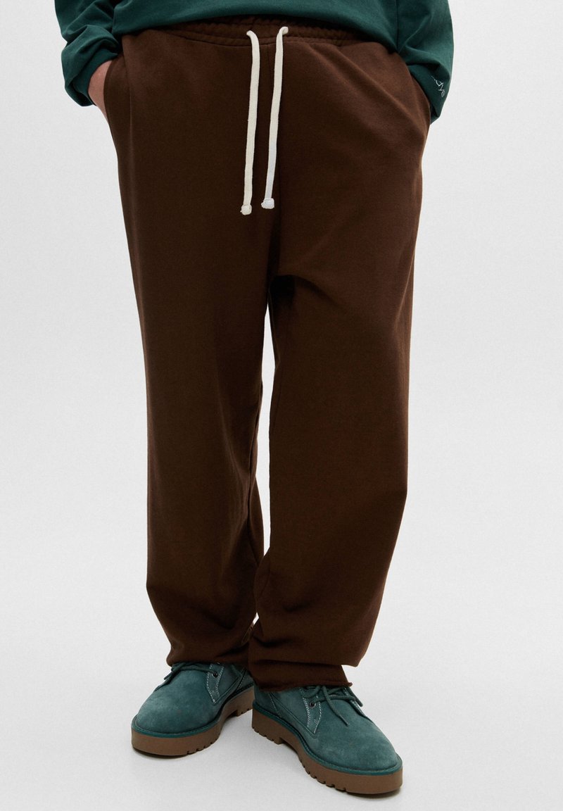 Brune sweatpants med elastisk talje og hvid snor, der har en afslappet pasform. Kombineret med tealfarvede sko og en grøn top.