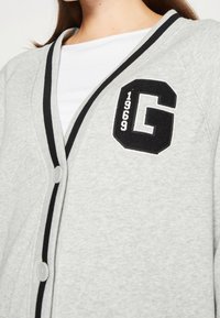 Cárdigan gris hecho de una mezcla de algodón, con ribetes negros, dos botones y un gran parche negro con la letra "G" y el año "1969".