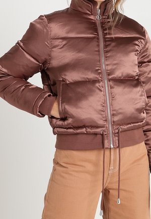 Femme portant une veste matelassée brillante marron avec fermeture éclair frontale, poignets élastiques et ourlet à cordon de serrage, associée à un pantalon marron clair.