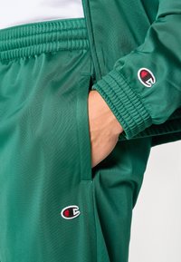 Πράσινο παντελόνι φόρμας από λεία ύφασμα με ελαστική μέση και τελειώματα με μανσέτες, με μ ικρό κεντημένο λογότυπο στην περιοχή της τσέπης.