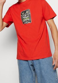 Camiseta de algodón roja con un gráfico de una cara distorsionada con dientes verdes. Mangas cortas y corte estándar. Jeans azules visibles por debajo.