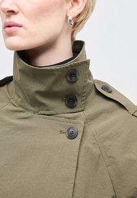 Κοντινό πλάνο ενός ατόμου που φοράει παλτό ελιάς με ψηλό γιακά κουμπωμένο με τρία σκούρα κουμπιά και ασημένιο σκουλαρίκι με κρίκο.