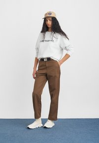 Szary sweter z logo "Carhartt", brązowe spodnie, beżowa czapka i białe trampki z czarnymi akcentami, noszone na jednolitym tle.