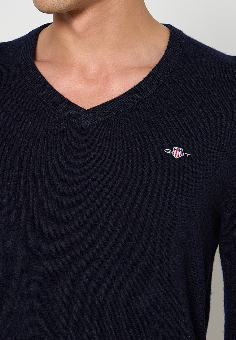 V-Ausschnitt Pullover in Marineblau, aus feinem Strickstoff gefertigt, mit kleinem besticktem Logo auf der Brust. Weicher Stoff und enganliegendes Design.