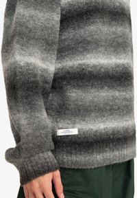 Grijs gestreepte trui gemaakt van dikke wol, met een ribbelmanchet en onderkant. Een wit label met blauwe tekst is zichtbaar aan de onderkant.