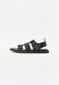 Sandales en cuir noir avec trois brides, une boucle sur le côté et une semelle en caoutchouc plate. Design simple avec peu de détails, sur fond blanc.