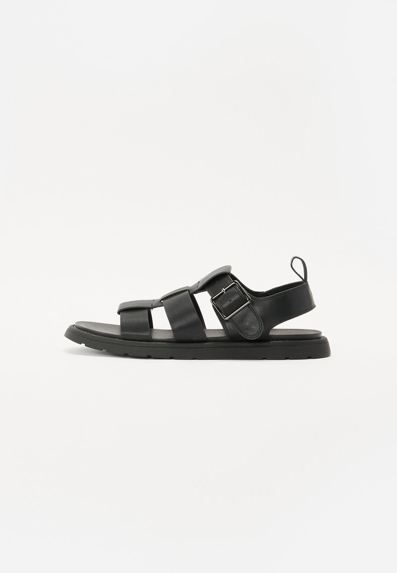 Sandales en cuir noir avec trois brides, une boucle sur le côté et une semelle en caoutchouc plate. Design simple avec peu de détails, sur fond blanc.