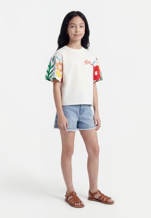 Κορίτσι στέκεται φορώντας άσπρο μπλουζάκι με πολύχρωμα μανίκια με floral σχέδιο, ανοιχτό μπλε σορτς από τζιν και καφέ σανδάλια με λουράκια σε λευκό φόντο.