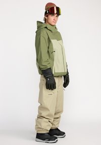Snowboarduitrusting met een groen en beige waterdichte jas, beige broeken, zwarte handschoenen en zwarte snowboardlaarzen met grijze accenten.