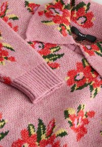 Gebreide pullover in zacht roze met een bloemenpatroon in rood, groen en geel. Heeft een kraag en geribde manchetten voor extra textuur.