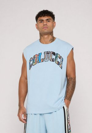 Mann med tatoveringer iført lys blå ermeløs skjorte og matchende shorts med fargerik "Colucci"-tekst på skjorten og bukselinjen.