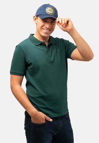 Camisa polo verde con cuello y mangas cortas, combinada con jeans de mezclilla oscuros. Se lleva una gorra azul con un emblema de árbol circular.