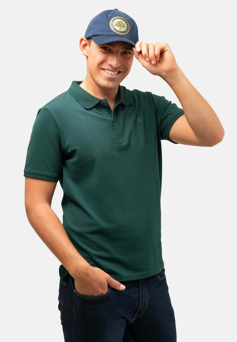 Camisa polo verde con cuello y mangas cortas, combinada con jeans de mezclilla oscuros. Se lleva una gorra azul con un emblema de árbol circular.