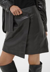 Falda de cuero negra con un diseño envolvente, que presenta un cierre de botón y una textura suave y brillante. El dobladillo es asimétrico, cayendo por encima de la rodilla.