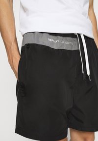 Shorts de bain noirs avec une ceinture élastique grise, dotés d'un cordon de serrage et d'un détail logo "REPLAY" en blanc. Fabriqués à partir d'un matériau lisse et léger.