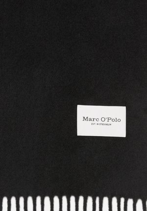 Tissu noir avec une étiquette rectangulaire blanche cousue, affichant le texte « Marc O’Polo EST. IN STOCKHOLM » et un bord inférieur effiloché.
