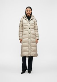 Beige polstret lang frakke med hætte, med trykknapper og en let finish, parret med mørke jeans og spidse sorte sko.