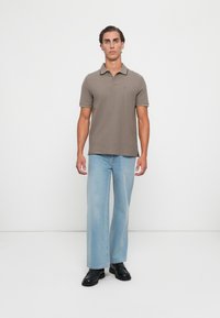 Mann som står mot hvit bakgrunn iført taupe kortermet polo, lyseblå vide jeans og svarte dressko.