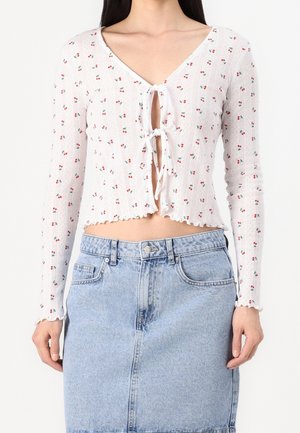 Crop top blanc à manches longues avec petit imprimé cerises rouges et fermetures à nouer devant, associé à une jupe en jean bleu clair.