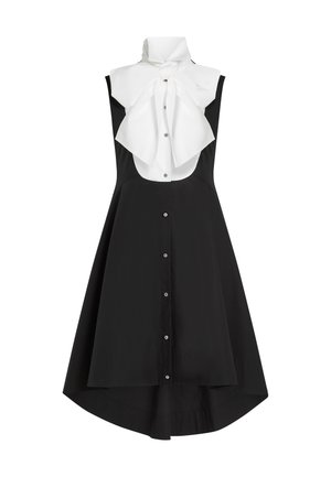 Robe noire sans manches avec ourlet asymétrique, patte de boutonnage à l'avant et col plastron blanc orné d'un grand nœud noué au niveau du cou.