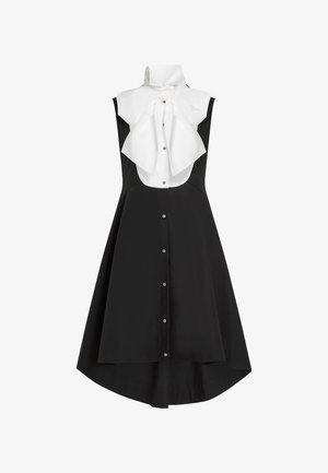 Mouwloze zwarte jurk met een high-low zoom, knoopsluiting aan de voorkant en een witte kraag met een grote, gestrikte strik bij de hals.