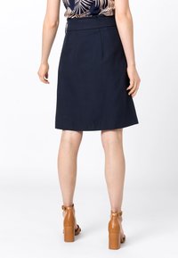 Jupe bleu marine avec une taille ajustée et des plis, longueur genou, faite d'un tissu texturé ; portée avec des sandales à talons carrés beiges.