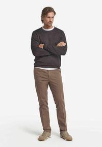 Mörk tröja med vit rundhals, bruna chinos och ljusa sneakers. Modellen står med armarna korslagda och visar upp outfitens passform och färgkontrast.