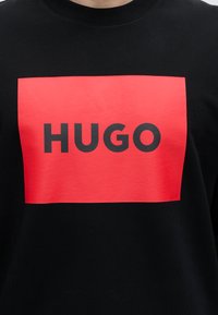 Must dressipluus suur punane ristküliku kujuline logo, millel on kiri „HUGO“ paksus mustas suurtes tähtedes.