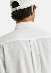 Weißes Leinenhemd mit einem Button-Down-Kragen und einem Rückenfalte-Detail. Der Stoff hat eine strukturierte, atmungsaktive Optik, die sich für warmes Wetter eignet.